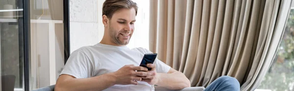 Mensajería hombre feliz en el teléfono inteligente mientras está sentado en casa cerca de cortina beige, pancarta - foto de stock