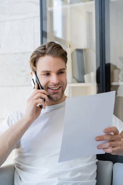 Jeune et heureux homme tenant du papier blanc pendant la conversation sur smartphone — Photo de stock