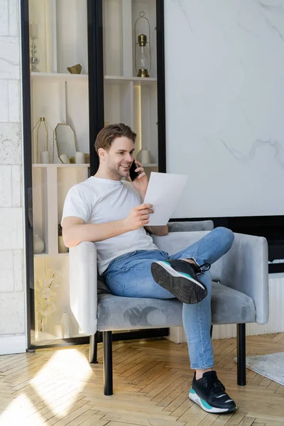 Visão de comprimento total do homem positivo com papel falando no celular na poltrona — Fotografia de Stock