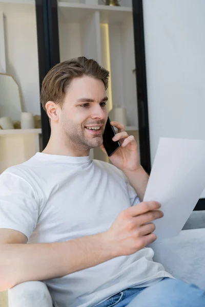 Улыбающийся мужчина держит бумагу и разговаривает по мобильному телефону дома — стоковое фото