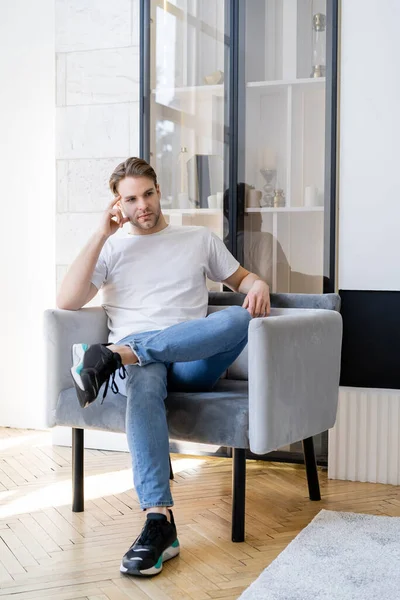 Nachdenklicher Mann in weißem T-Shirt sitzt mit überkreuzten Beinen im Sessel und schaut weg — Stockfoto