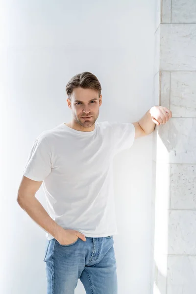 Hombre en camiseta blanca de pie con la mano en el bolsillo de jeans cerca de la pared blanca - foto de stock