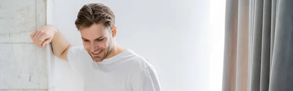 Uomo sorridente in t-shirt bianca in piedi vicino al muro bianco a casa, banner — Foto stock