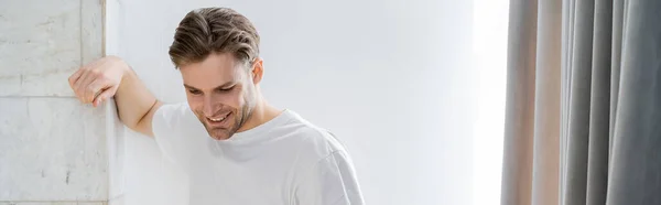 Homem sorridente em t-shirt branca de pé perto da parede branca em casa, banner — Fotografia de Stock