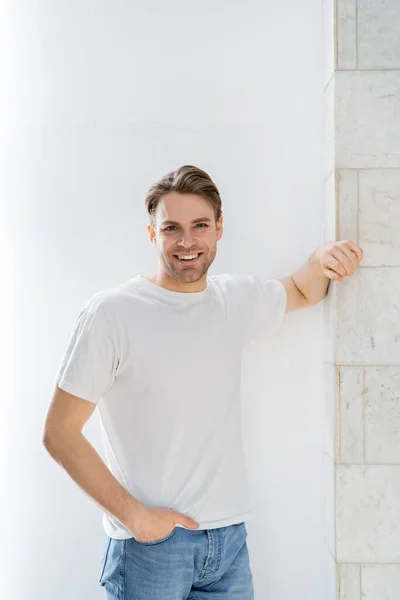 Jovem e feliz homem de pé com a mão no bolso de jeans perto da parede branca — Fotografia de Stock