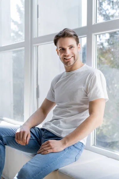 Homem feliz em t-shirt branca sentado no peitoril da janela em casa — Fotografia de Stock