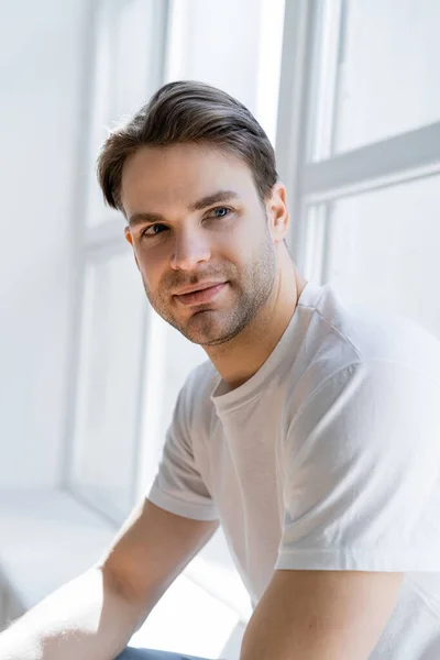 Porträt eines positiven Mannes im weißen T-Shirt, der in der Nähe des Fensters zu Hause wegschaut — Stockfoto