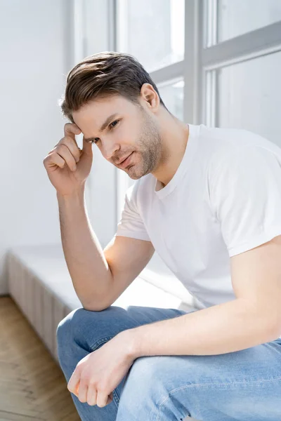 Positive and dreamy man holding hand near head while sitting on windowsill — Stock Photo