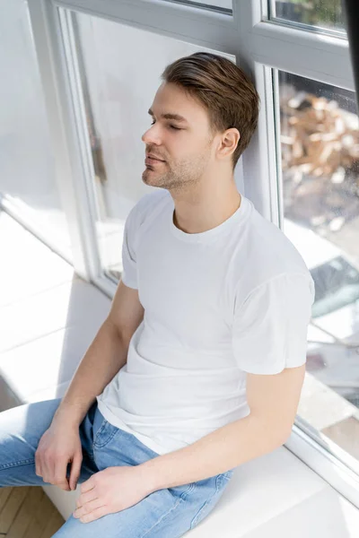 Молодой человек в белой футболке сидит на подоконнике с закрытыми глазами — стоковое фото