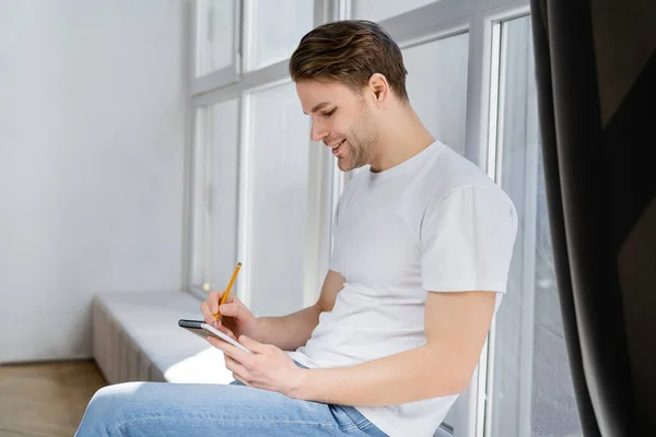 Vista lateral del hombre satisfecho sentado en el alféizar de la ventana y la escritura en el cuaderno - foto de stock