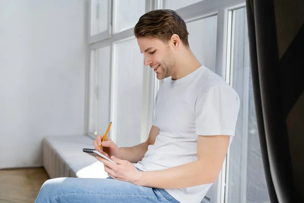 Vue de côté de l'homme heureux assis sur le rebord de la fenêtre et l'écriture dans le carnet — Photo de stock