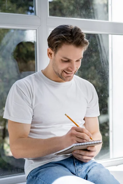 Uomo sorridente che scrive nel diario vicino alla finestra di casa — Foto stock