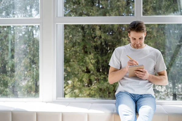 Joven en camiseta blanca y jeans sentado en el alféizar de la ventana y la escritura en el diario - foto de stock