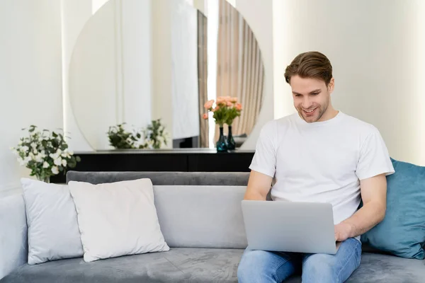 Hombre feliz sentado en la sala de estar decorado con flores y trabajando en el ordenador portátil - foto de stock