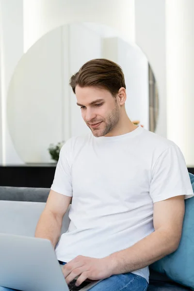 Jovem e sorridente homem em branco t-shirt trabalhando no laptop em casa — Fotografia de Stock