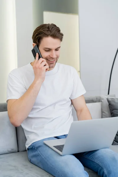 Hombre alegre que llama en el teléfono móvil mientras trabaja en el ordenador portátil en casa - foto de stock