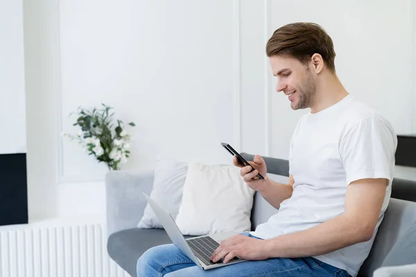 Vue latérale de l'homme souriant assis avec smartphone et ordinateur portable sur le canapé à la maison — Photo de stock