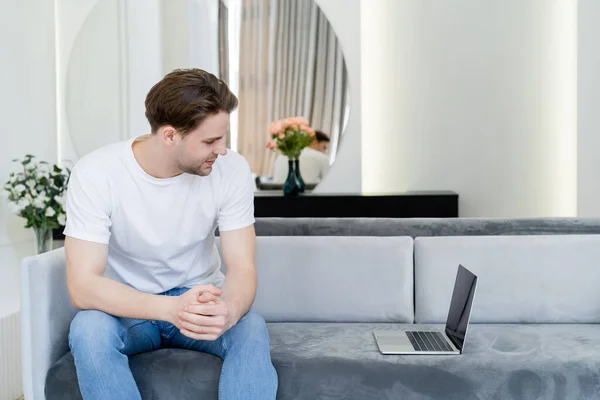 Young man sitting on sofa in living room and looking at laptop with blank screen — Stock Photo