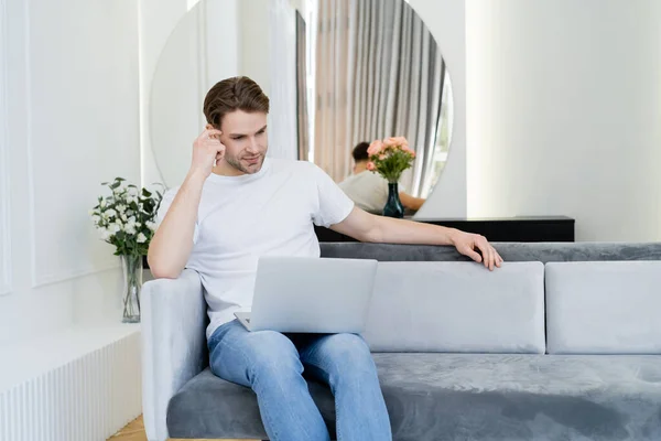 Nachdenklicher junger Mann sitzt auf Sofa in der Nähe von Blumenvasen und schaut auf Laptop — Stockfoto