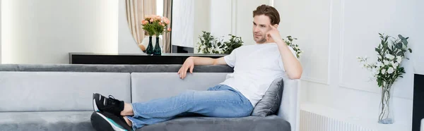Vue pleine longueur de l'homme réfléchi assis sur le canapé dans le salon avec décoration florale, bannière — Photo de stock