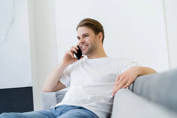Cheerful young man sitting on blurred couch and talking on mobile phone — Stock Photo