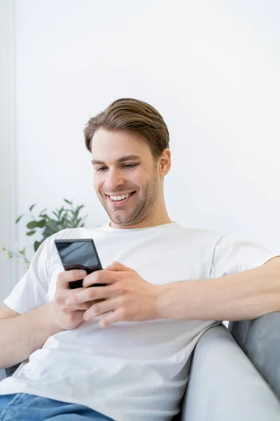 Alegre joven en blanco camiseta mensajería en el teléfono celular mientras está sentado en casa - foto de stock