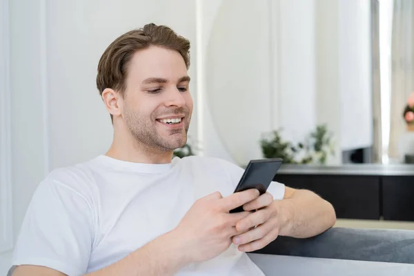 Щасливий молодий чоловік у білій футболці, використовуючи мобільний телефон вдома — стокове фото