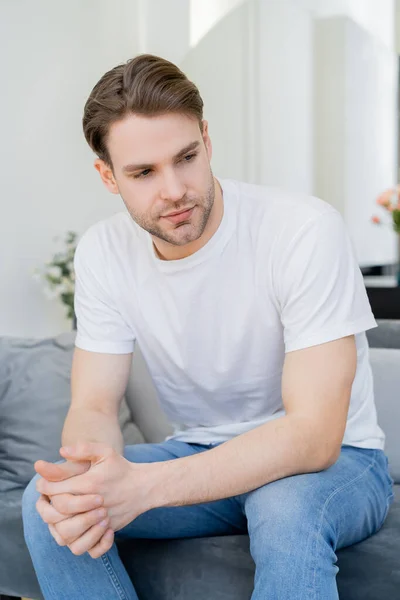 Jovem homem em t-shirt branca sentado com as mãos apertadas na sala de estar — Fotografia de Stock