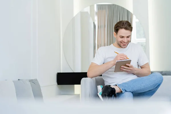 Cheerful man writing in notebook while sitting in living room on blurred foreground — Stock Photo