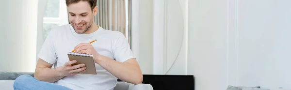 Hombre positivo en camiseta blanca escribiendo en cuaderno mientras está sentado en casa, pancarta - foto de stock
