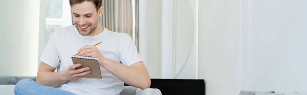 Positive man in white t-shirt writing in notebook while sitting at home, banner — Stock Photo