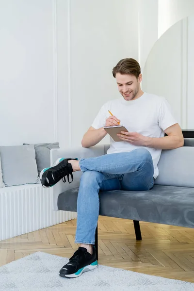 Ganzkörperansicht eines lächelnden Mannes, der mit überkreuzten Beinen sitzt und in ein Notizbuch schreibt — Stockfoto