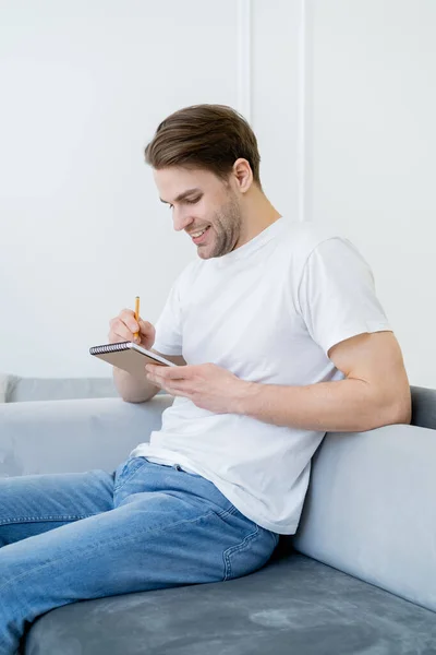 Щасливий чоловік у білій футболці пише в блокноті, сидячи на дивані вдома — стокове фото