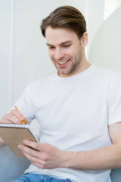 Молодой и счастливый мужчина в белой футболке, пишущий в дневнике дома — стоковое фото