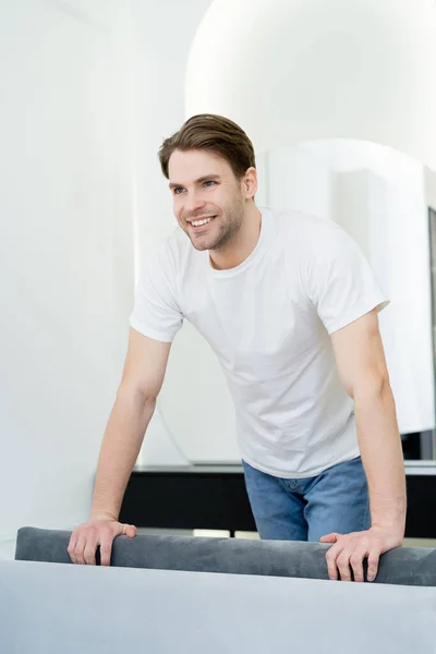 Junger Mann im weißen T-Shirt lächelt, während er im Wohnzimmer steht — Stockfoto