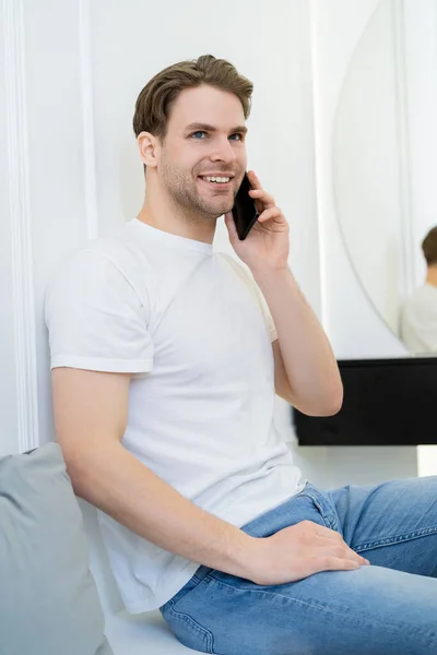 Uomo sorridente guardando lontano mentre seduto sul divano e parlando sul telefono cellulare — Foto stock