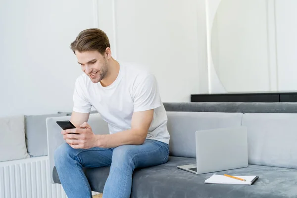 Uomo sorridente seduto sul divano vicino al computer portatile e messaggistica su smartphone — Foto stock