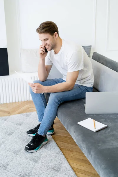 Homme souriant regardant le document tout en appelant sur smartphone près d'un ordinateur portable — Photo de stock