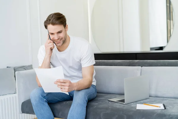Positive man talking on cellphone while looking at document near laptop on couch — Stock Photo