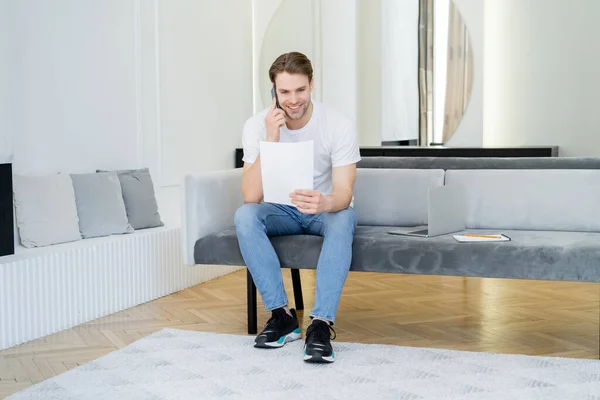 Vista completa del hombre con el papel hablando en el teléfono móvil cerca del ordenador portátil en el sofá - foto de stock