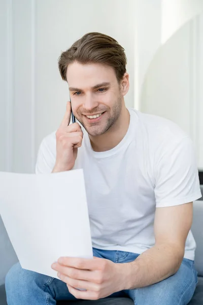 Smiling man in white t-shirt looking at paper while talking on cellphone — Stock Photo