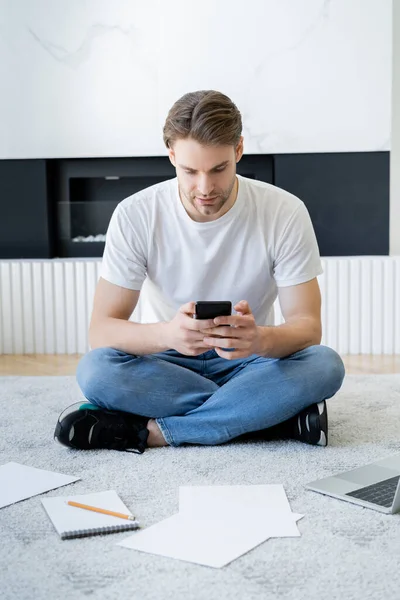 Hombre sentado en el suelo con las piernas cruzadas y la mensajería en el teléfono inteligente cerca de la computadora portátil y papeles - foto de stock