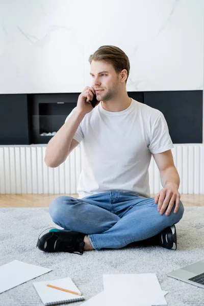 Freelancer sitting on floor with crossed legs, looking away and talking on smartphone — Stock Photo