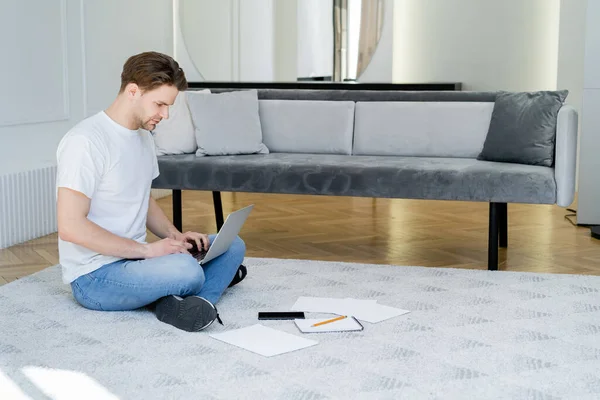 Comprimento total do homem sentado no chão na sala de estar e digitando no laptop — Fotografia de Stock