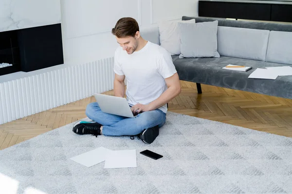 Smiling man typing on laptop while sitting on floor near papers and smartphone with blank screen — Stock Photo