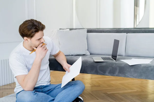 Hombre reflexivo con lápiz y papel sentado con las piernas cruzadas cerca del ordenador portátil con pantalla en blanco - foto de stock