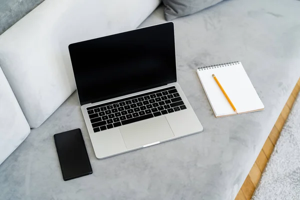 Portátil y teléfono móvil con pantalla en blanco cerca del cuaderno y lápiz en el sofá gris - foto de stock