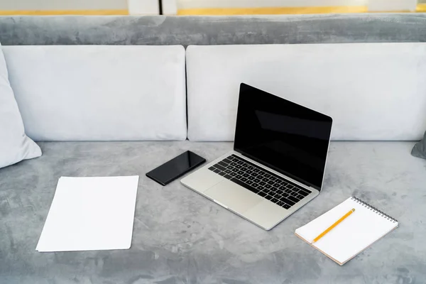 Papeles vacíos, cuaderno y lápiz cerca de la computadora portátil y el teléfono inteligente con pantalla en blanco en el sofá gris - foto de stock