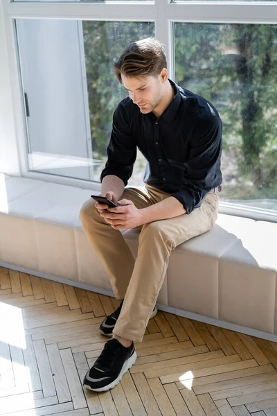 Mann in schwarzem Hemd und schwarzer Hose auf Fensterbank sitzend und Nachrichten auf Smartphone — Stockfoto