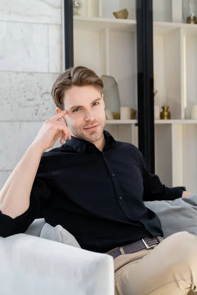 Joven hombre con estilo en camisa negra sentado en sillón y mirando a la cámara - foto de stock